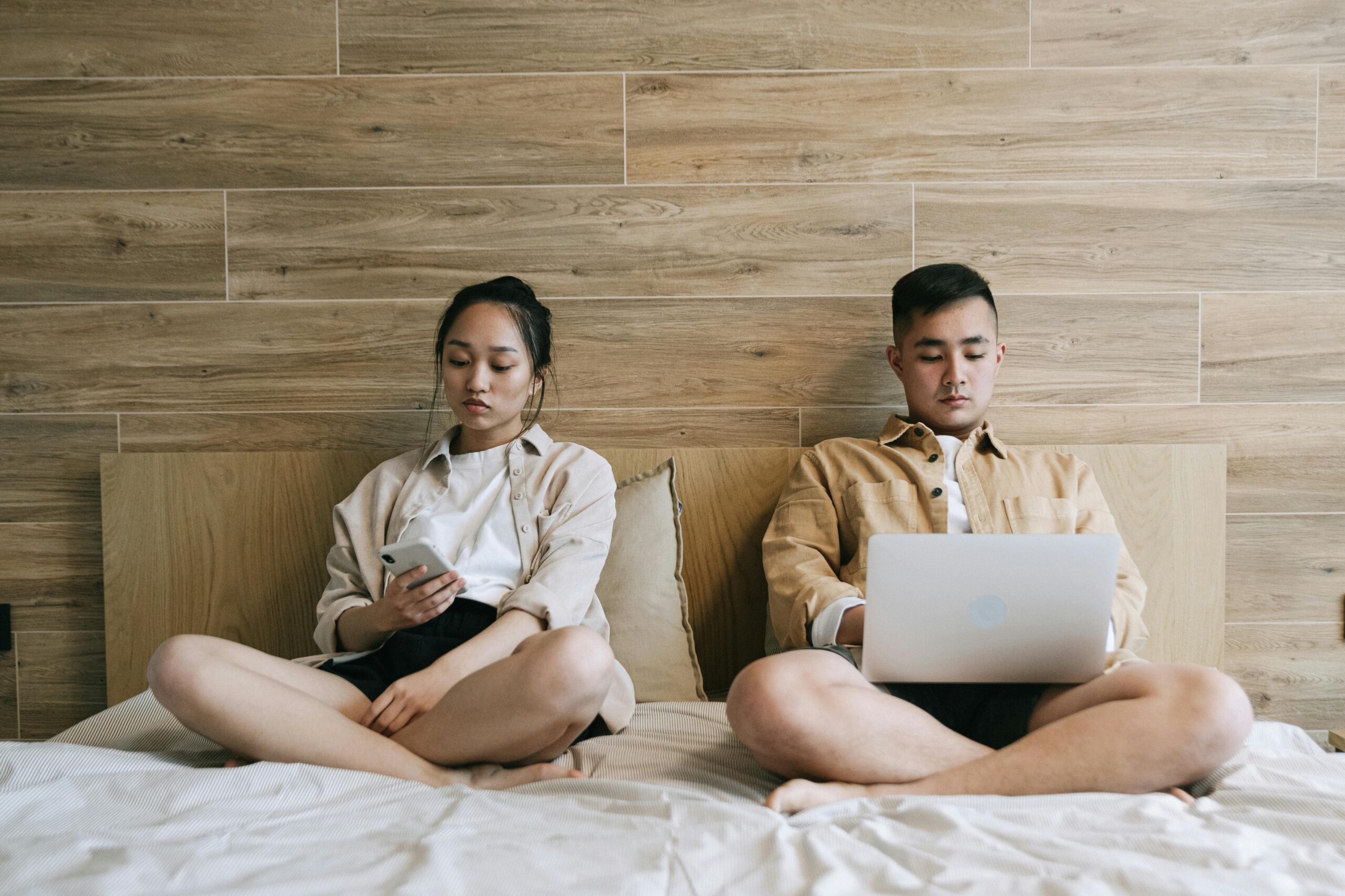 A young couple sits apart on a bed against a wooden wall, both looking down at their devices—a phone and a laptop—appearing emotionally distant after a conflict.