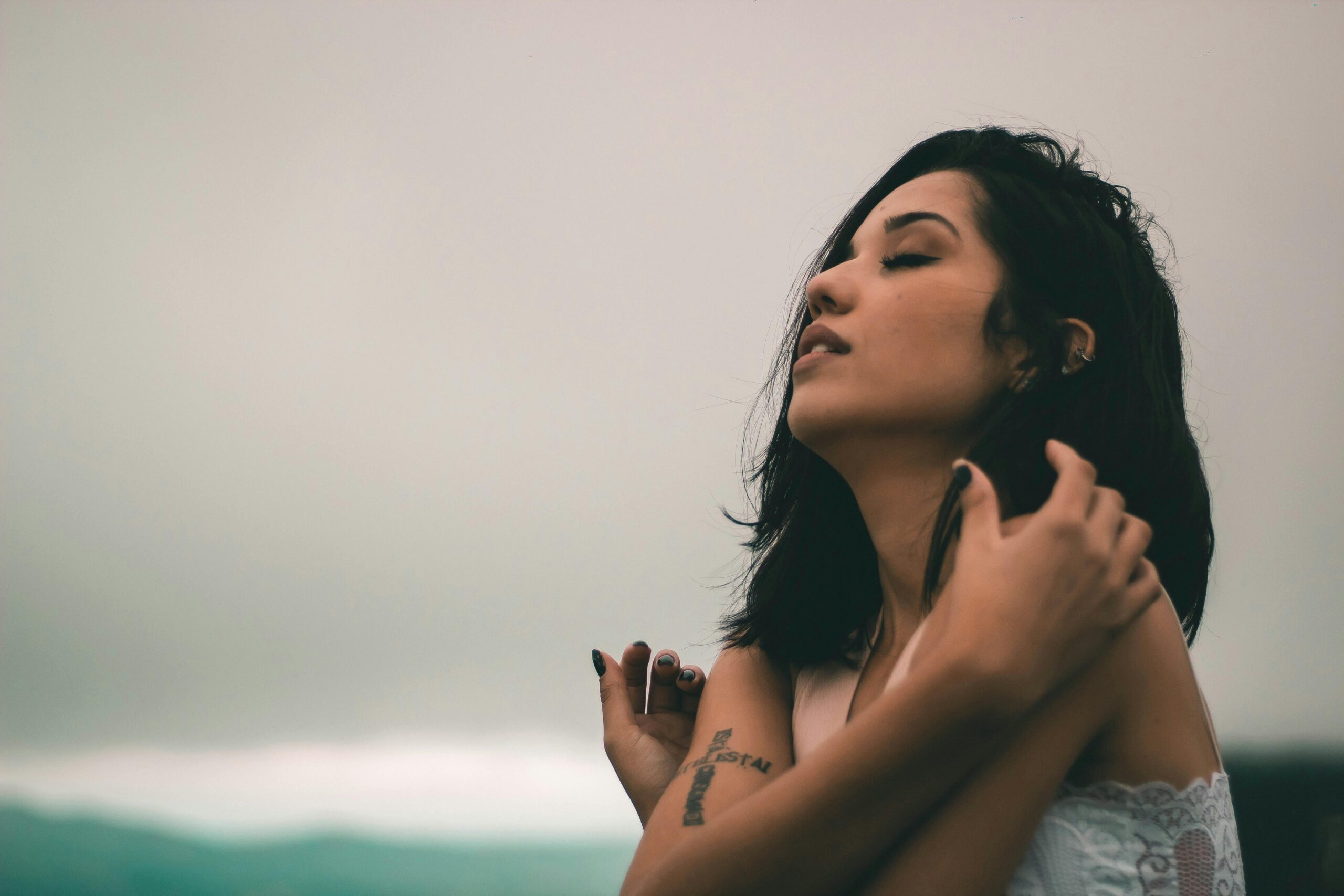 Side-profile of a woman with eyes closed, gently brushing her hair back outdoors against a soft, cloudy sky and blurred ocean horizon.