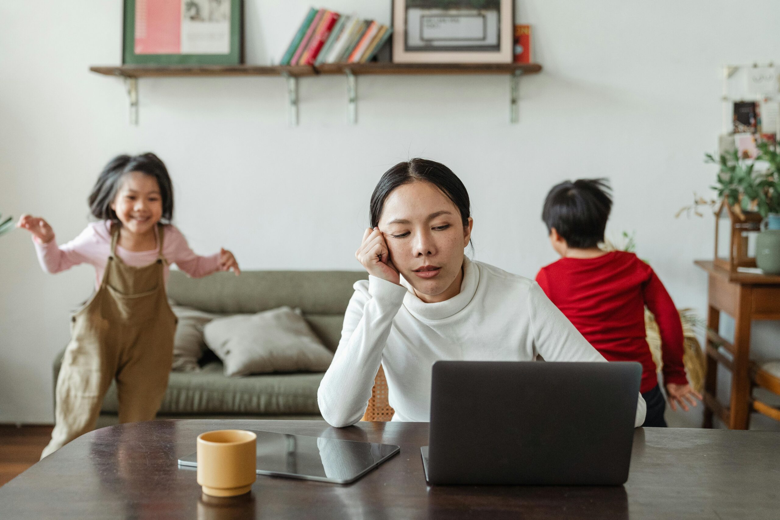 A woman sits at a dining table working on a laptop with her head resting on her hand, looking tired, while two young children play and move around in the background of a cozy living room.