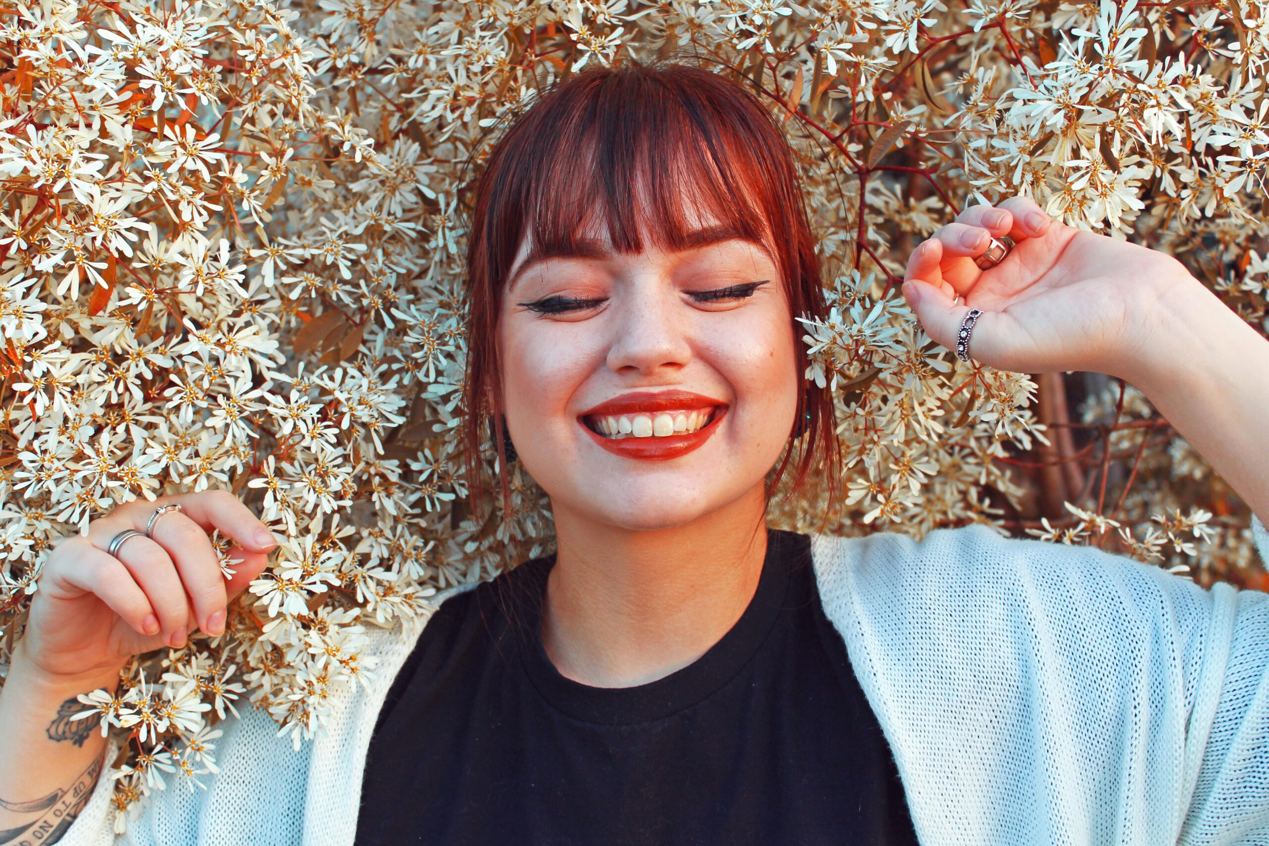 Smiling woman with red lipstick and bangs stands among white flowering branches with her eyes closed.