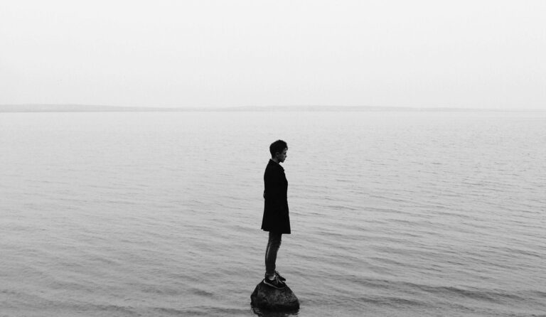 Black-and-white photo of a solitary person standing on a rock in calm, open water, looking down toward the surface, with a faint horizon line in the distance, evoking reflection and emotional weight.