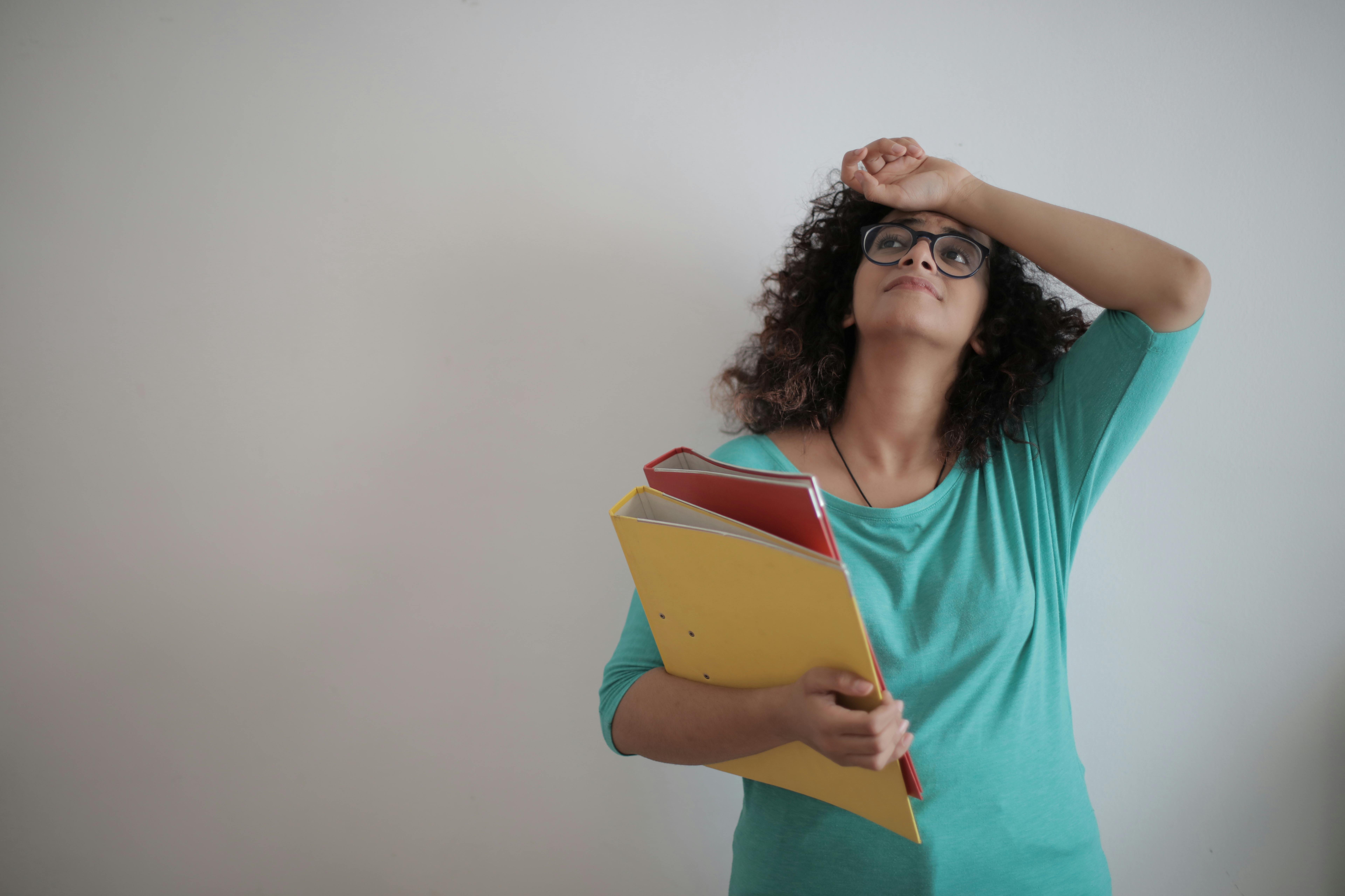 Person with curly hair and glasses holding colorful folders, looking upward with a hand on their forehead against a plain wall, conveying stress or overwhelm.
