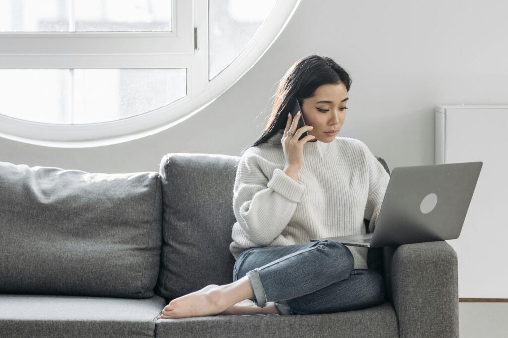 Person sitting cross-legged on a gray couch, holding a phone to their ear while working on a laptop near a large round window.