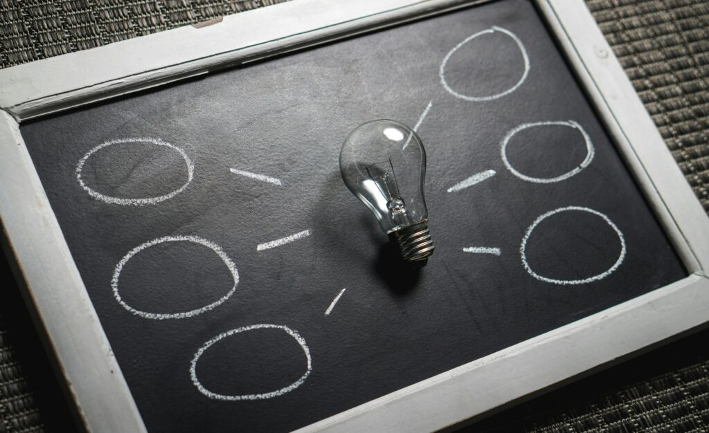 A light bulb rests on a small chalkboard surrounded by chalk-drawn circles connected by short lines, like a simple mind map or thought bubbles.