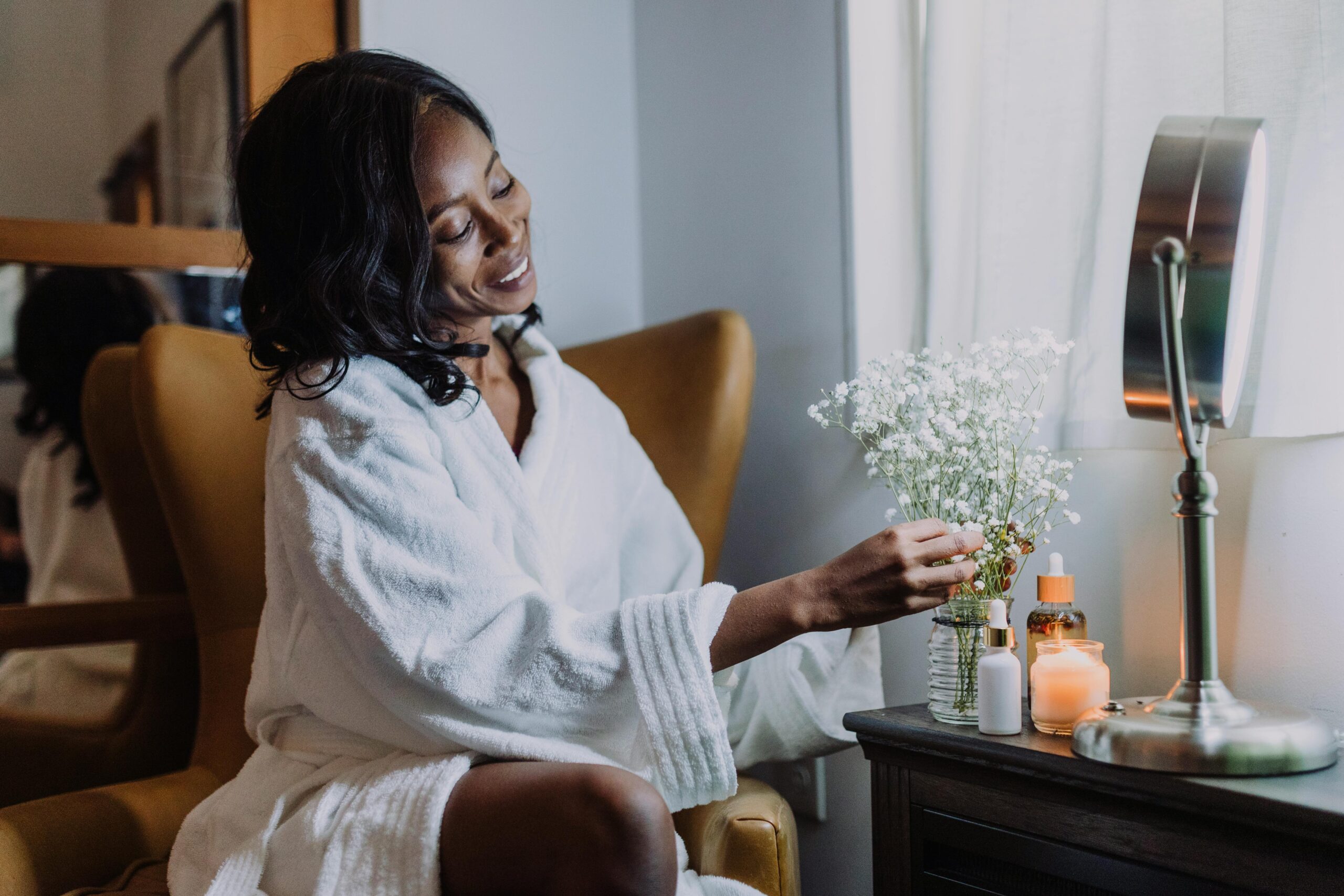 Woman in a white robe sitting by a window, gently arranging small white flowers on a vanity table with a mirror, candles, and skincare bottles, creating a calm and cozy self-care setting.