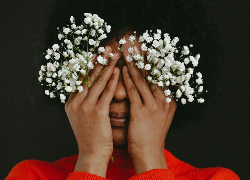 Close-up portrait of a person in a red sweater covering their eyes with their hands while holding small white flowers (baby’s breath) against a dark background.
