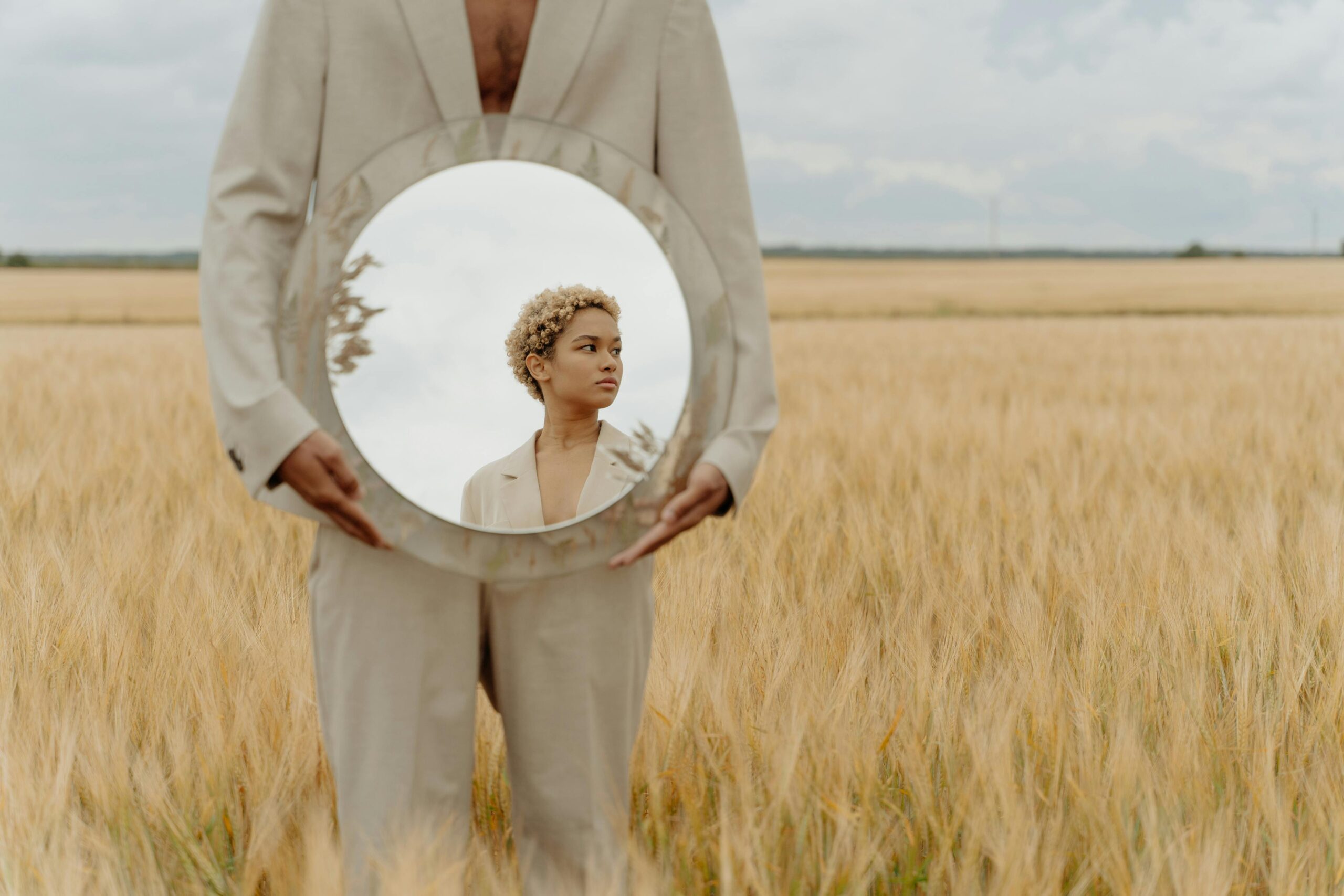 Person standing in a golden wheat field holding a round mirror that reflects the face of a thoughtful young woman in a beige blazer, symbolizing identity and self-reflection beyond one’s past.