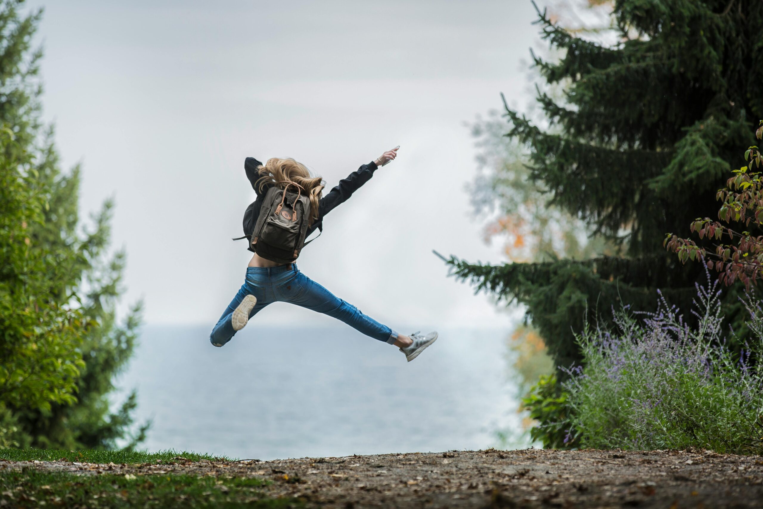 Person with a backpack jumping joyfully on a forest trail overlooking a lake, symbolizing renewed energy and overcoming seasonal energy slumps.