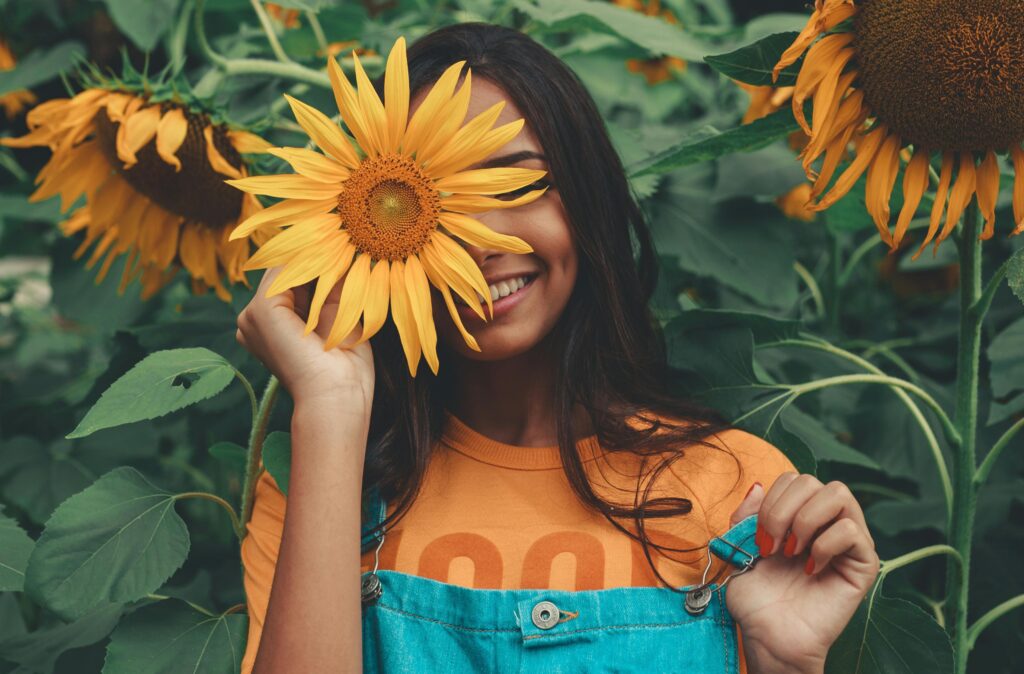 Smiling woman holding a sunflower over one eye while standing in a sunflower field, symbolizing authenticity, self-expression, and being real in therapy for mental health and personal growth.